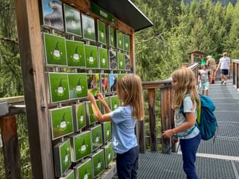 Two girls playing an interactive memory game on a wooden treetop walkway in Saalbach-Hinterglemm. Green panels with white symbols mounted on display.