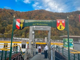 Ferry dock with green archway labeled 'Engelhartszell I - Auf Wiedersehen' with coats of arms. Yellow and white passenger ships docked on the Danube.