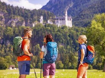 Drei Wanderer mit Rucksäcken machen Pause auf einer Wiese und betrachten Schloss Neuschwanstein in der Ferne, umgeben von bewaldeten Bergen.