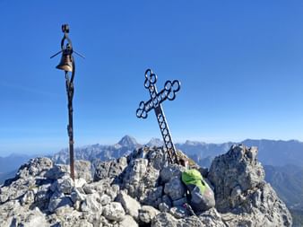 Metallenes Gipfelkreuz und Glocke auf felsigem Gipfel des Cima del Cacciatore mit Wanderer in grüner Jacke und Rucksack vor Bergpanorama.