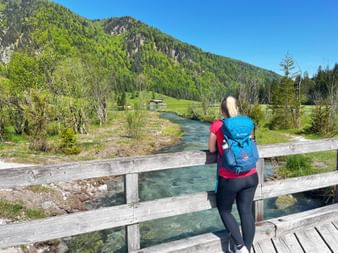 Female hiker with blue backpack on wooden bridge over turquoise river near Pillersee, forested mountains and meadows in background.