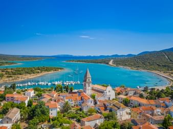 Aerial view of Osor, a coastal town with red-roofed houses and a church tower, overlooking a turquoise bay with sailboats in Kvarner Bay, Croatia.
