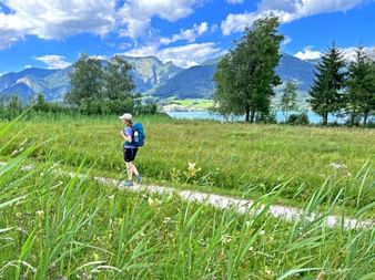 Hiker with blue backpack walking on grassy path beside turquoise lake in Salzkammergut, with mountains and trees under blue sky.