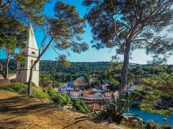 View of Veli Lošinj village with colorful houses and church tower, framed by pine trees. Forested hills and blue water visible in background.