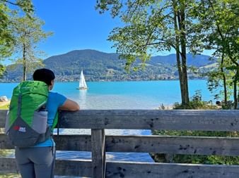 Wanderin in grüner Jacke an Holzbank mit Blick auf den Tegernsee mit Segelboot, Bergen und Bäumen, die das türkisfarbene Wasser umrahmen.