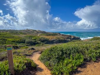 Hölzerner Wegweiserpfosten am Küstenpfad bei Praia das Furnas mit grüner Vegetation, Meersklippen und blauem Himmel mit weißen Wolken.