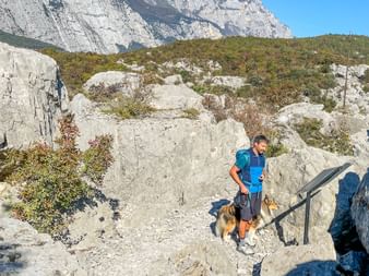 Männlicher Wanderer mit blauem Rucksack und seinem Hund zwischen großen Kalksteinfelsen in bergigem Gelände mit Vegetation und steilen Klippen.
