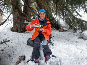 Lächelnder Wanderer in oranger Jacke und blauer Mütze sitzt im Schnee unter Nadelbaum und gießt Getränk aus Thermoskanne in Becher.