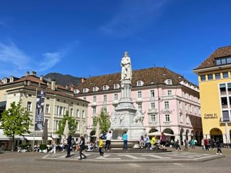 Walther Square in Bozen with white monument statue in center, surrounded by colorful historic buildings and people walking across the plaza.