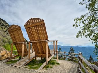 Two oversized wooden chairs at Trauttmansdorffer Thronsessel viewpoint on Meraner Höhenweg, overlooking mountain valleys and peaks.