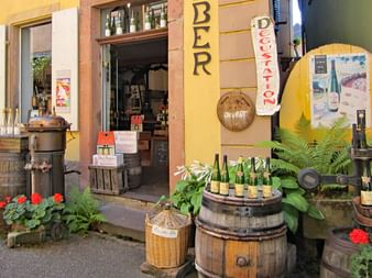 Bunter Weinladen-Eingang im Elsass mit Holzfässern, Weinflaschen und 'BIER'- und 'DÉGUSTATION'-Schildern an gelber Fassade.