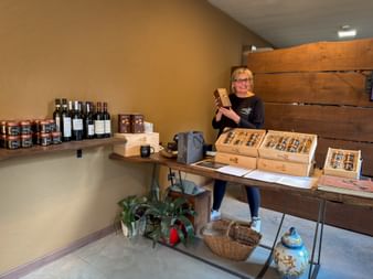 Woman with glasses in delicatessen in Cravanzana, Piedmont, standing behind table with wooden boxes of products, wine bottles and jars on shelves.
