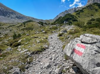 Rot-weiße Wegmarkierung auf Felsen entlang eines steinigen Bergpfads im Karwendel bei Innsbruck, mit grünen Almwiesen und Gipfeln.