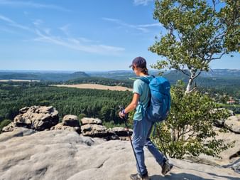 Wanderer mit blauem Rucksack auf felsigem Aussichtspunkt am Gohrisch mit Blick auf bewaldete Täler und Berge unter blauem Himmel.