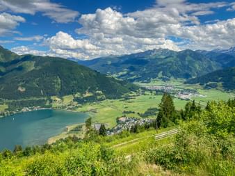 Panoramic view from Schmittenhöhe over Zeller See lake and Zell am See town, surrounded by green meadows and forested mountains under blue sky.