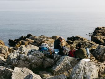 Zwei Personen sitzen auf großen Küstenfelsen bei Stenshuvud und picknicken am ruhigen Meer unter bedecktem Himmel.