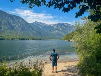 Hiker with backpack standing on a pebble beach at Lake Bohinj, facing mountains of the Julian Alps under blue sky with scattered clouds.