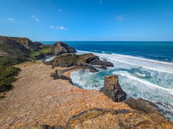 Rocky cliffs at Praia da Pena Furada with turquoise waves crashing against rock formations. Green vegetation covers the clifftops under blue sky.