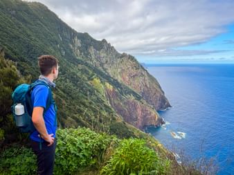 Hiker with blue backpack on Vereda do Larano trail overlooking steep green cliffs and deep blue Atlantic Ocean on Madeira's north coast.
