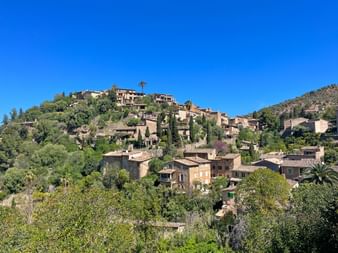 Stone houses of Deia village nestled on a green hillside in Mallorca, surrounded by trees and vegetation under a clear blue sky.