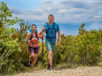 Zwei Wanderer mit Rucksäcken und Wanderstöcken auf einem Schotterweg, umgeben von grünen Büschen unter blauem Himmel mit Bergen im Hintergrund.