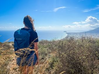 Female hiker with blue backpack standing on Monte Piccaro overlooking the Ligurian coast and sea with coastal town visible below.