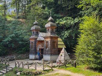 Hölzerne russische Kapelle mit zwei Zwiebeltürmen in einer Waldlichtung, umgeben von einem Holzzaun. Zwei Besucher stehen am Eingang.