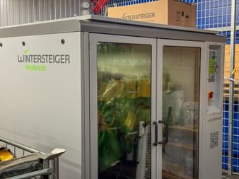 White Wintersteiger Velobrush automated bike washing station with glass doors showing green and yellow cleaning brushes inside.
