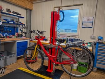 Red bicycle mounted on repair stand in workshop with blue tool cabinets, workbench, and window showing outdoor view in Schärding.