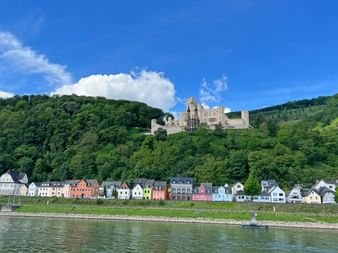 Schloss Stolzenfels thront auf einem bewaldeten Hang über bunten Häusern am Rhein. Blauer Himmel mit weißen Wolken über grünen Hügeln.
