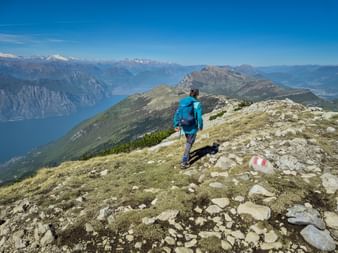 Hiker in blue jacket walking on Monte Baldo ridge trail with Lake Garda and snow-capped Alps visible in the background under clear blue sky.
