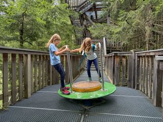 Two girls balancing on a colorful rotating play platform on an elevated treetop walkway in Saalbach-Hinterglemm, Pinzgau.