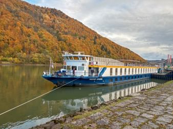 Blue and white river cruise ship moored at cobblestone dock on the Danube. Hillside with autumn foliage in orange and yellow tones rises behind.