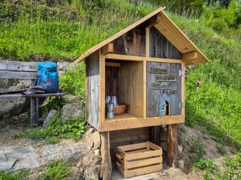 Wooden self-service stand on Meraner Höhenweg trail with lemon balm juice bottles and basket inside. A blue backpack rests on a bench nearby.
