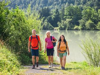 Three hikers with backpacks walking on a path beside the Drau River in Carinthia, surrounded by lush green forest and hills.