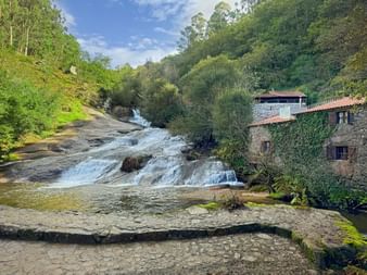 Fervenzas do Barosa Wasserfälle fließen über Felsen neben einer Steinmühle mit rotem Ziegeldach, umgeben von üppigem grünen Wald.
