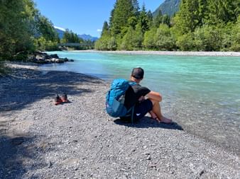 Wanderer mit blauem Rucksack sitzt am Kiesufer des türkisfarbenen Lech, umgeben von grünem Wald und Bergen unter blauem Himmel.