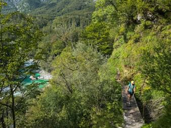 Wanderin mit Rucksack auf schmalem Bergweg über dem türkisfarbenen Fluss Soča in den Julischen Alpen, umgeben von üppiger grüner Vegetation.