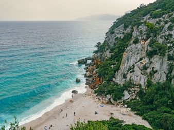 Aerial view of Cala Fuili beach with turquoise water, white pebble shore, and steep rocky cliffs covered in green vegetation on Sardinia's coast.