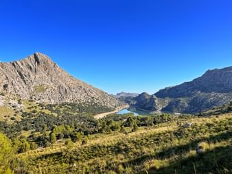 Gorg Blau reservoir nestled between rocky Tramuntana mountains with green vegetation in foreground and clear blue sky above.