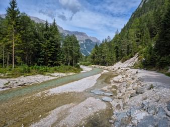 Leutascher Ache mit klarem Wasser fließt durch ein felsiges Tal nahe der Zugspitze, umgeben von dichtem Wald und Bergen unter blauem Himmel.
