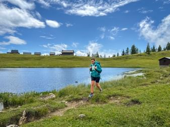 Wanderin mit Rucksack steht am Tauplitzsee auf der Tauplitzalm. Almhütten und Nadelbäume auf grünen Hügeln unter blauem Himmel sichtbar.