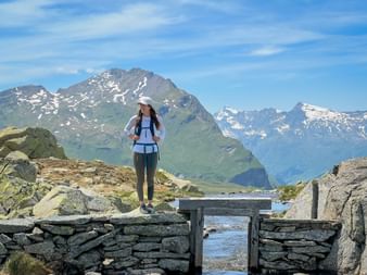 Wanderin auf Holzbrücke über Bergsee Surettasee mit schneebedeckten Alpengipfeln im Hintergrund unter blauem Himmel auf der Via Spluga.