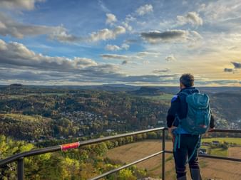 Hiker with blue backpack on Lilienstein viewing platform overlooking forested valley and villages under dramatic cloudy sky on Malerweg trail.