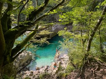 Türkisfarbener Soča-Fluss mit felsigem Ufer und Steinbrücke, eingerahmt von moosbewachsenen Bäumen. Zwei Personen auf Felsen am Wasser.