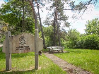 Hölzerne Infotafel mit Ammoniten-Logo und Aufschrift 'Naturpark Altmühltal' auf grüner Wiese mit Bank und Bäumen im Hintergrund.