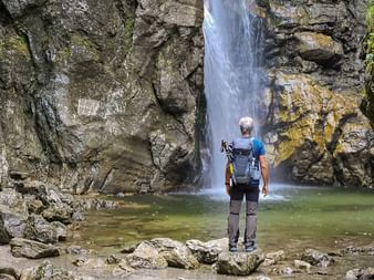 Hiker with backpack and camera standing on rocks before a waterfall cascading into a pool, surrounded by moss-covered rock walls.
