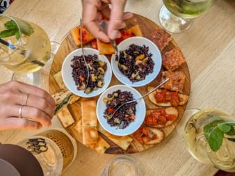 Wooden board with three bowls of black rice salad, bruschetta, and crackers. Hands reaching for food, white wine glasses visible around the table.