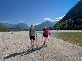 Two women hiking with poles on gravel riverbank of Soča River in Julian Alps. Mountains and forest visible under blue sky.