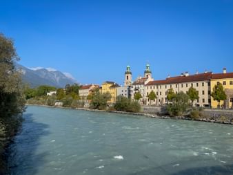 Türkisfarbener Inn fließt durch Innsbruck mit bunten historischen Gebäuden und Kirchtürmen am Flussufer, Berge im Hintergrund.